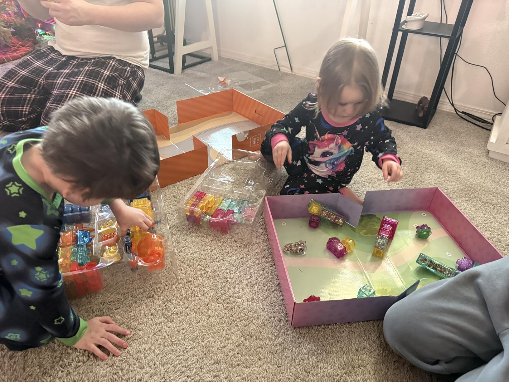 Two kids in pajamas opening a Construction Site box and a pink castle set on the living room floor